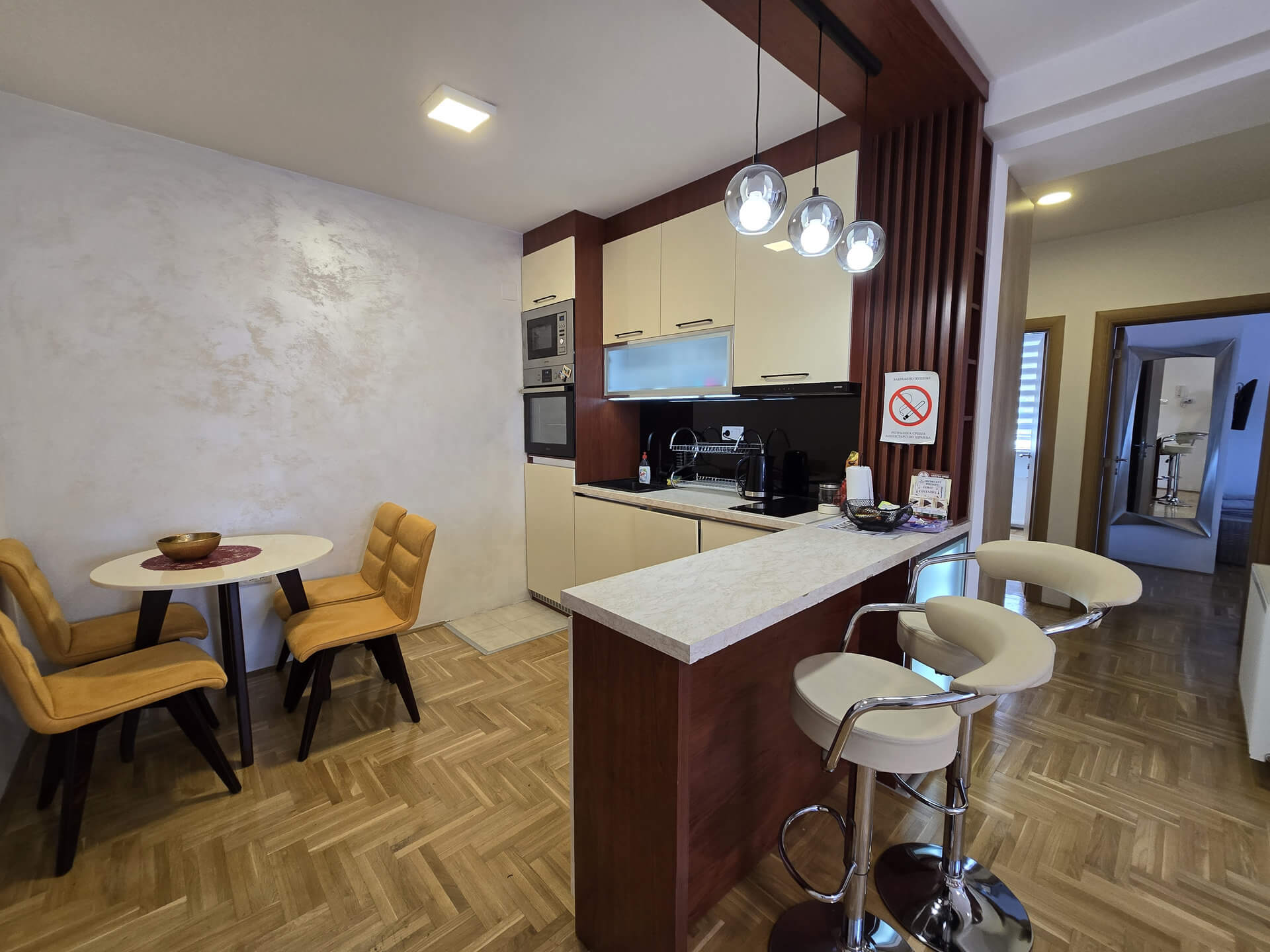 Kuhinja sa trpezarijom, šankić sa dve barske stolice, mali sto u levom uglu, lampe vise iznad šanka, kuhinja sa ugradnom mašinom za sudove i mikrotalasnom